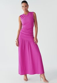 A model poses confidently in a vibrant pink sleeveless dress, accessorized with gold jewelry and nude heels, against a neutral background.