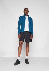 Chaqueta de forro polar verde azulado con cuello alto, camiseta blanca, pantalones cortos cargo negros y zapatos negros con calcetines blancos. El modelo está de pie sobre un fondo liso.