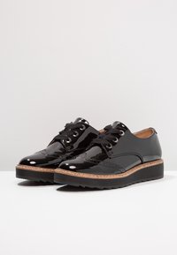 Chaussures en cuir verni noir avec un détail brogue, bout rond et lacets noirs ; elles présentent une semelle intermédiaire en liège et une semelle en caoutchouc texturé.