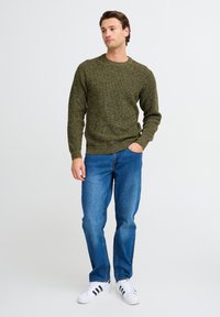 Olijfgroene, gebreide trui met een ronde hals, gecombineerd met blauwe jeans en witte sneakers met zwarte strepen. Klassieke casual outfit.