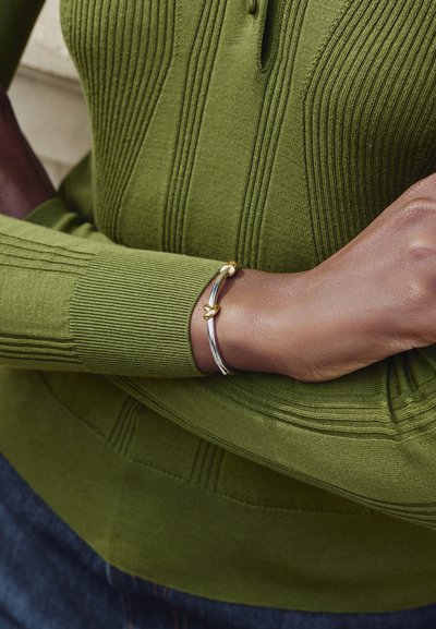 Κοντινό πλάνο βραχίονα σταυρωμένου πάνω στον κορμό, που φοράει μια ριγωτή ελιά πράσινη μπλούζα και ασημένιο βραχιόλι με χρυσές λεπτομέρειες κόμπου.