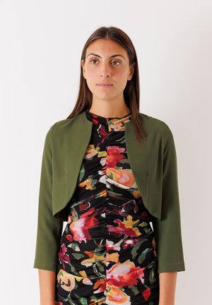 Giacca cropped verde oliva con maniche a tre quarti sopra un vestito aderente floreale con design arricciato in colori vivaci su uno sfondo nero.