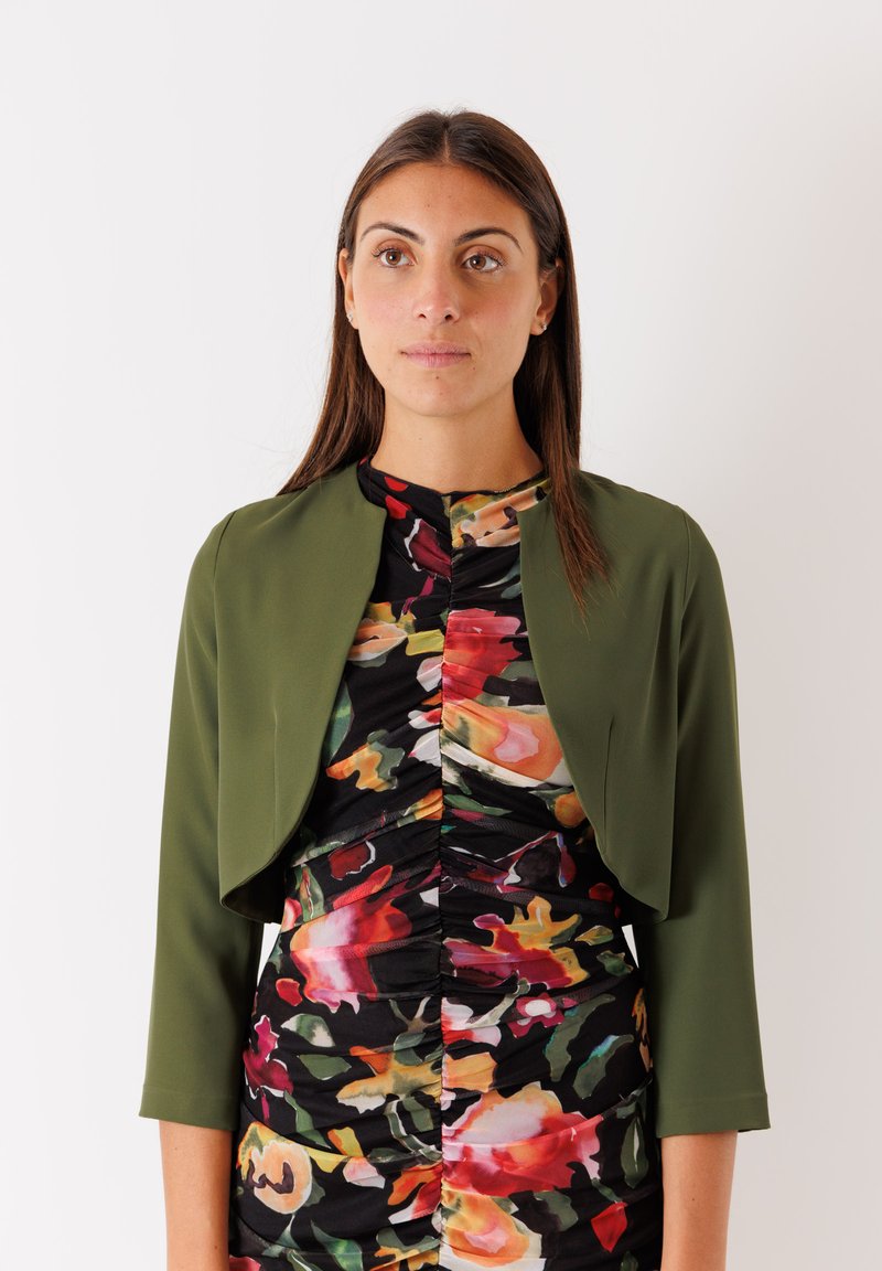 Giacca cropped verde oliva con maniche a tre quarti sopra un vestito aderente floreale con design arricciato in colori vivaci su uno sfondo nero.