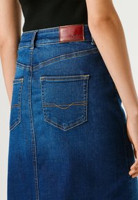 Inimene kannab sinist teksaseelikut tagataskutega ja pruuni nahast brändi plaastriga, mida on kombineeritud musta topiga.