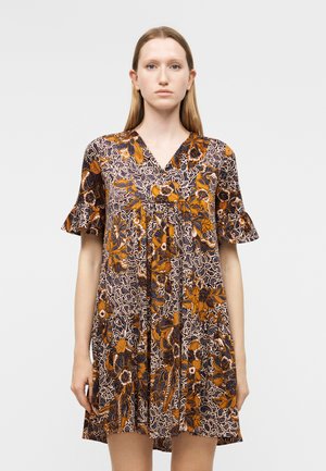 Jauna moteris dėvi trumpomis rankovėmis gėlėtą suknelę oranžinės, juodos ir baltos spalvų raštais, stovinti priešais paprastą šviesiai pilką foną.