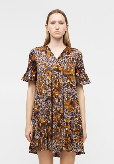 Jeune femme portant une robe courte à manches courtes à motif floral orange, noir et blanc, posant devant un fond uni gris clair.