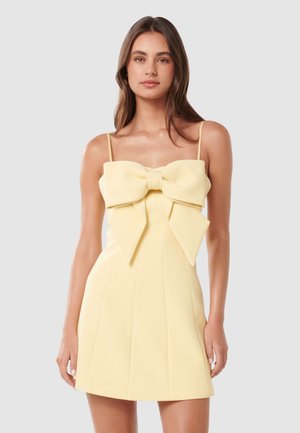 Žena nosi bledozlté mini šaty bez rukávov s tenkými ramienkami a veľkým mašľovým detailom na hrudi na jednoduchom pozadí.