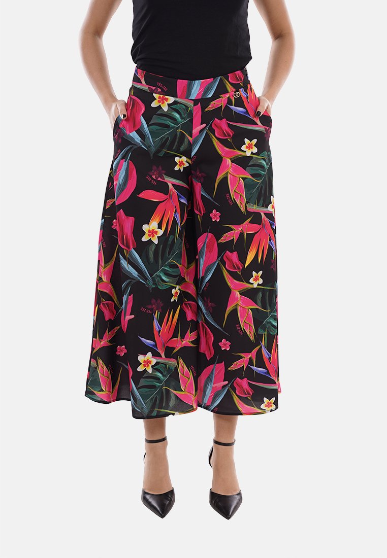 Donna che indossa pantaloni neri a gamba larga cropped con vivace stampa floreale tropicale e tacchi neri con cinghia alla caviglia, mani in tasca, in piedi.