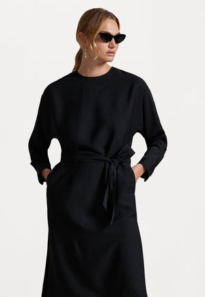 Musta pitkähihaista mekkoa, jossa on pyöreä kaula-aukko, kudottu pinta, vyöllinen vyötärö ja sivutaskut. Minimalistinen muotoilu puhtailla linjoilla.