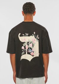Black cotton t-shirt featuring a large floral design with pink flowers and green leaves overlaying a stylized letter on the back.