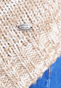 Beżowa dziergana czapka z teksturowanym wzorem z grubej przędzy. Metalowa metka z napisem "KHUJO EST. 2001 ROUGH & FINE." Niebieska tkanina widoczna poniżej.