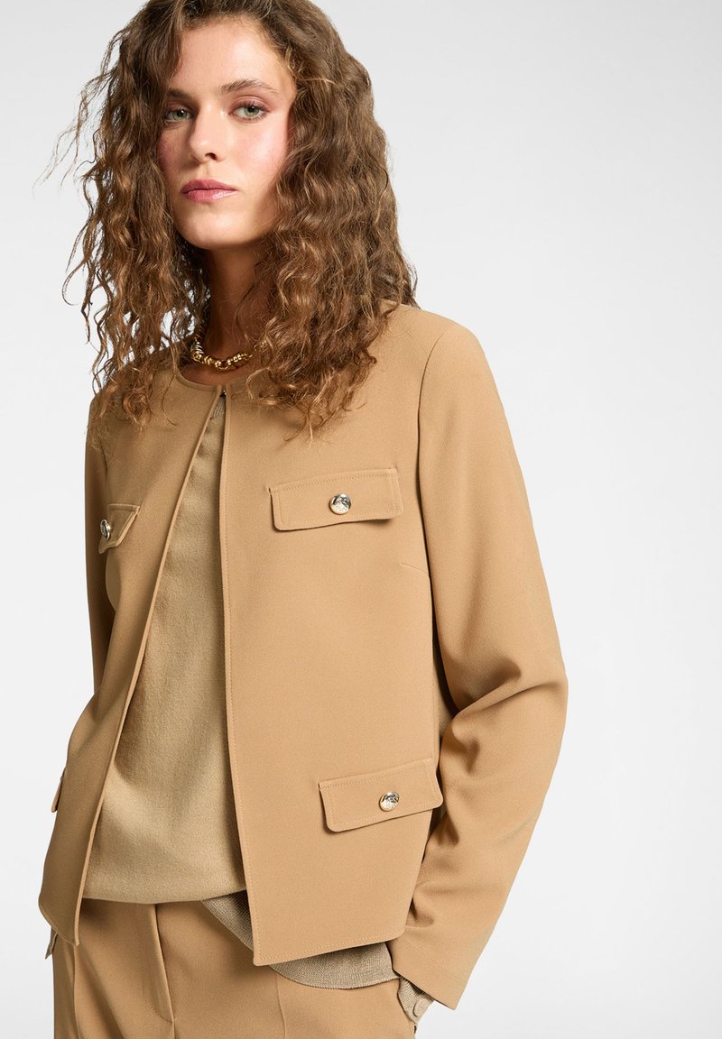 Béžový blazer s kulatým výstřihem, dvě přední kapsy se zlatými knoflíky, hladký materiál, nošený přes světle béžový top. Vyznačuje se měkkým, přizpůsobeným střihem.