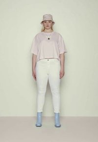 Top de manga curta rosa claro, jeans justos brancos e botins azuis. O modelo usa um chapéu de balde estampado e colares em camadas.