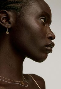 Pendiente de aro dorado con un diseño de estallido solar, combinado con delicadas cadenas de oro y plata, exhibido sobre piel oscura y un fondo neutro.