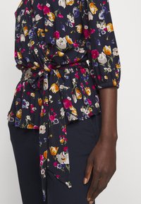 Blommig blus med marinblå bakgrund, prydd med flerfärgade blomster mönster, markerad midja med knyt detalj, veckade ärmar och lätt tyg.