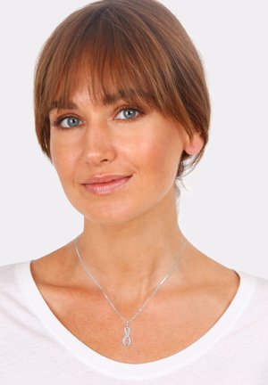 Collana d'argento con pendente a simbolo dell'infinito, adornata da una delicata catena, indossata sopra una camicia bianca. Design semplice ed elegante.