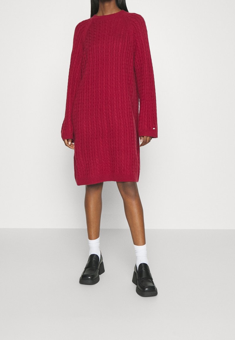 Femme portant une robe-pull rouge torsadée mi-longue, à manches longues, avec des chaussettes blanches et des mocassins noirs épais.