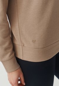 Bež dukserica s okruglim rubom, teksturiranom tkaninom i šivenim detaljima. Ima diskretni logo na donjoj lijevoj strani.