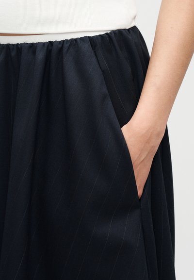 Χέρι στην τσέπη μαύρης φούστας με λεπτές ρίγες και ελαστική μέση, σε συνδυασμό με λευκό τοπ, πάνω σε απλό φόντο.