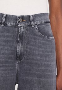 Taille et partie supérieure de la cuisse d'une personne portant un jean en denim noir délavé à taille haute avec un bouton en métal et un haut blanc.