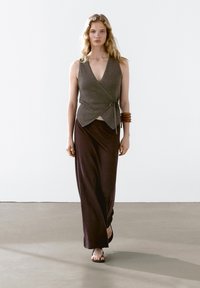 Grijze, wikkelstijl top gecombineerd met een vloeiende, donkerbruine rok. Gladde gebreide textuur met een casual ontwerp. G gedragen met zwarte sandalen en een bruine armband.
