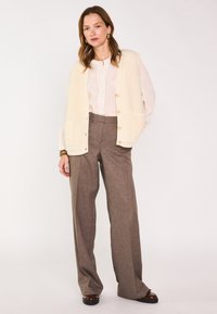 Gilet en tricot ivoire avec des boutons, associé à un pantalon à carreaux brun taille haute et une blouse crème avec des détails froncés.