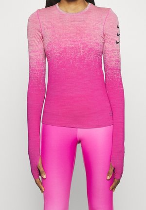 Femme portant un haut de sport manches longues rose ombré avec des trous pour les pouces et des leggings rose vif assortis, debout sur un fond blanc.