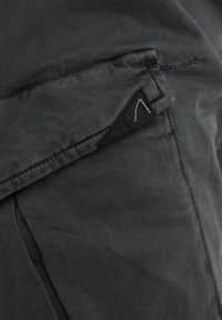 Gros plan sur une poche en tissu gris foncé avec rabat et coutures visibles, montrant une petite étiquette en tissu noir avec un détail en fil blanc.