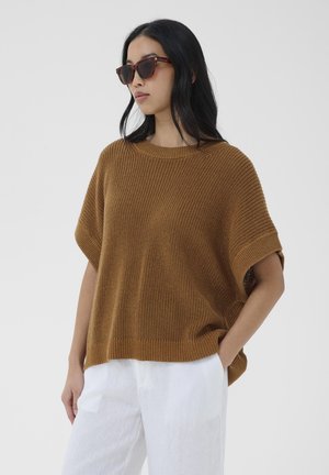 Femme portant des lunettes de soleil écaille, un pull en tricot marron ample et un pantalon blanc, debout avec une main dans la poche sur fond gris.