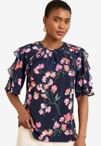 Blusa floreale con sfondo blu navy, caratterizzata da fiori rosa e verdi. Le maniche arricciate e il collo rotondo aggiungono texture e dettagli.
