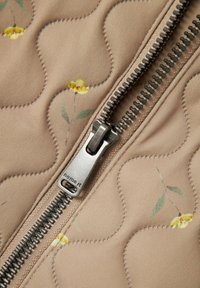 Quiltet stof i blød beige med små gule blomstermønstre, med en sølvmetal lynlås langs en kant.