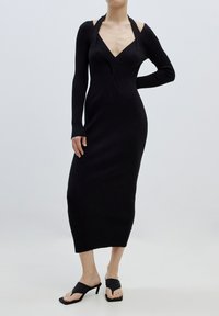 Schwarzes, geripptes Strickkleid mit langen Ärmeln und tiefem V-Ausschnitt sowie taillierter Silhouette, ausgestattet mit ausgeschnittenen Schulterdetails. Ein Paar schwarze, hochhackige Sandalen.