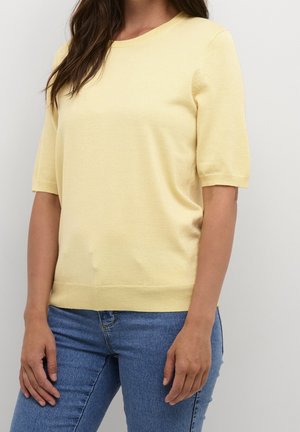 Femme portant un pull léger à manches courtes jaune pâle et un jean en denim bleu, debout devant un fond clair uni.