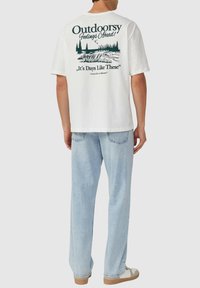 Witte katoenen t-shirt met een groene grafische afbeelding op de achterkant in outdoor-thema, met bomen, water en tekst. Gecombineerd met lichtblauwe jeans en casual schoenen.