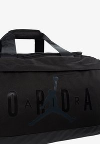 Bolsa de deporte negra hecha de una tela duradera, con una textura suave, logotipo en relieve y asas de transporte resistentes. Cierre de cremallera en la parte superior.