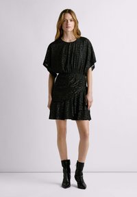 Robe noire à manches courtes et taille froncée, avec un motif texturé et un ourlet à volants. Associée à des bottines noires.