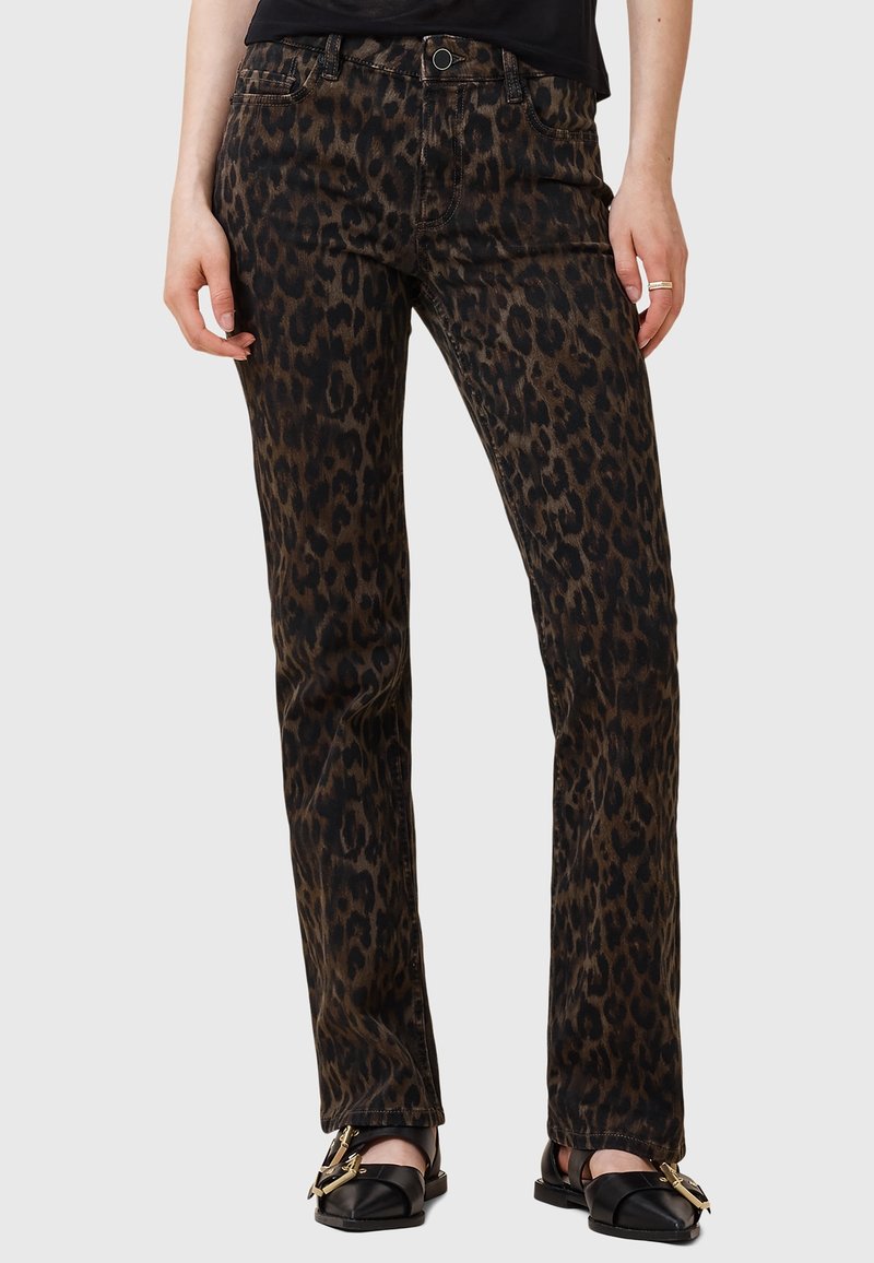 Femme portant un pantalon droit imprimé léopard marron foncé avec des ballerines noires à boucle dorée sur fond uni.