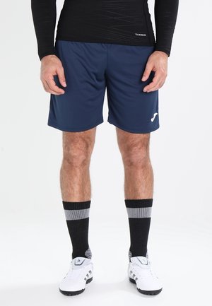 Mann trägt marineblaue Sportshorts, schwarz-graue wadenlange Socken, weiße Sportschuhe und ein schwarzes langärmliges Oberteil, steht vor weißem Hintergrund.