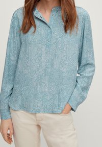 Frau in einem hellblauen gemusterten Langarmshirt mit Druckknöpfen und beigen Hosen, mit einer Hand in ihrer Tasche.