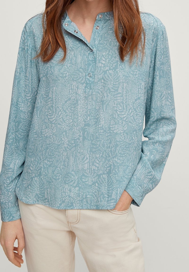 Frau in einem hellblauen gemusterten Langarmshirt mit Druckknöpfen und beigen Hosen, mit einer Hand in ihrer Tasche.