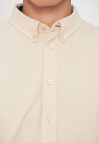 Chemise beige en velours côtelé à boutons avec col boutonné portée par une personne, montrant le cou et le haut de la poitrine.