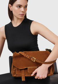 Borsa a tracolla in suede marrone con una tracolla regolabile, chiusura a patta e tasca frontale. Superficie texturizzata con dettagli in metallo dorato.