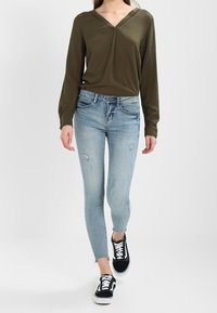 Olivgrön långärmad blus med V-ringning, i kombination med ljusblå slitna skinny jeans och svarta sneakers.