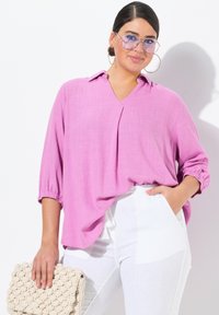 Blusa rosa, ampia e morbida, con scollatura a V e maniche di tre quarti. Abbinata a pantaloni bianchi, tenuta con una borsa beige a struttura.