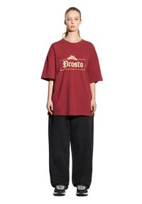 Burgundowa, oversize'owa koszulka z wyraźnym logo "Prosto" w kremowym kolorze, zestawiona z luźnymi czarnymi spodniami i sportowymi butami z szarymi akcentami.