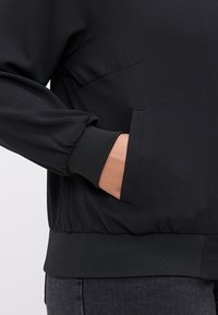 Black jacket with textured fabric, ribbed cuffs, and waistband. Features a side pocket with a sleek design and a relaxed fit.