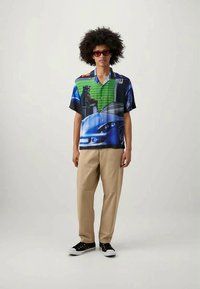 Young man wearing sunglasses, colorful graphic short-sleeve shirt with car and dog print, beige pants, black sneakers, standing against a plain background.