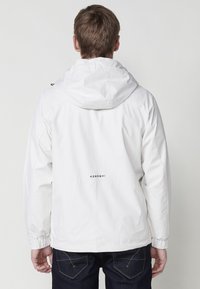Witte waterdichte jas met capuchon, elastische manchetten en een logo op de rug. Gladde textuur, minimalistisch ontwerp en ontspannen pasvorm.