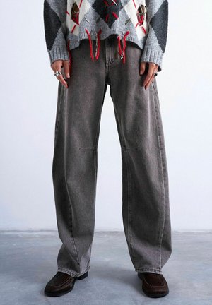 Personne portant un jean gris ample associé à des chaussures marron foncé et un pull gris à motifs avec des accents rouges et des ourlets usés.