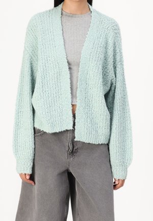 Kobieta ubrana w jasnoniebieski teksturowany sweter na guziki, na szary prążkowany crop top, do tego luźne szare jeansy z wysokim stanem.